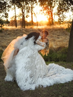 A couple is sharing an intimate moment in a forest setting during sunset. The woman is wearing an elaborate, feathered wedding dress, and the man is in a formal suit. They are surrounded by tall trees and dry grass, with the warm glow of the sun in the background creating a romantic atmosphere.