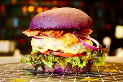 A gourmet burger with a vibrant purple bun, melted cheese, crispy bacon, slices of red onion, tomato, and fresh green lettuce. The burger is placed on a patterned surface with a blurred background featuring warm, ambient lighting.