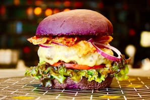 A gourmet burger with a vibrant purple bun, melted cheese, crispy bacon, slices of red onion, tomato, and fresh green lettuce. The burger is placed on a patterned surface with a blurred background featuring warm, ambient lighting.