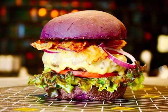 A gourmet burger with a vibrant purple bun, melted cheese, crispy bacon, slices of red onion, tomato, and fresh green lettuce. The burger is placed on a patterned surface with a blurred background featuring warm, ambient lighting.