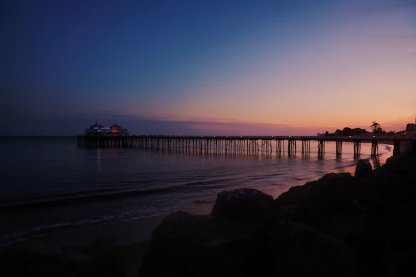 A wide shot of a wooden pier extending into calm coastal waters under a terracotta sunset.