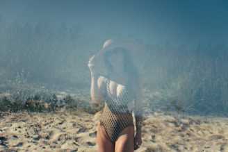 A person wearing a patterned swimsuit and a large sun hat stands on a sandy beach. The background features a hazy, blurred view of foliage, giving the setting a dreamy, vintage atmosphere.
