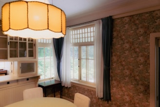 A cozy interior room with floral wallpaper and large windows letting in natural light. A round dining table with white chairs sits beneath a large, warm-toned pendant lamp. Blue and white curtains frame the windows, and a built-in cabinet with frosted glass adds a vintage charm.
