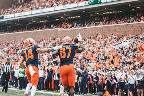 Two football players in navy and orange uniforms celebrate a touchdown in front of a large, cheering crowd. One referee and other team members can be seen in the background, with the brick stadium providing a classic sports venue backdrop.