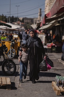 A woman in dark clothing and a headscarf walks arm in arm with a young boy through a busy market area. She carries several black plastic bags. Various market stalls, a yellow motorbike, and blurred figures of other people and distant buildings are visible in the background.