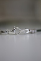 Close-up view of a metallic jewelry clasp on a blurred background. The clasp is in focus and detailed, showing its smooth, shiny surface and mechanism for opening and closing.