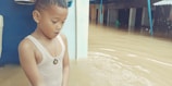 A young child stands in floodwater up to their waist, wearing a sleeveless shirt and necklace. The child looks concerned or surprised. In the background, partially submerged structures and objects are visible.