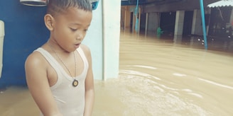 A young child stands in floodwater up to their waist, wearing a sleeveless shirt and necklace. The child looks concerned or surprised. In the background, partially submerged structures and objects are visible.
