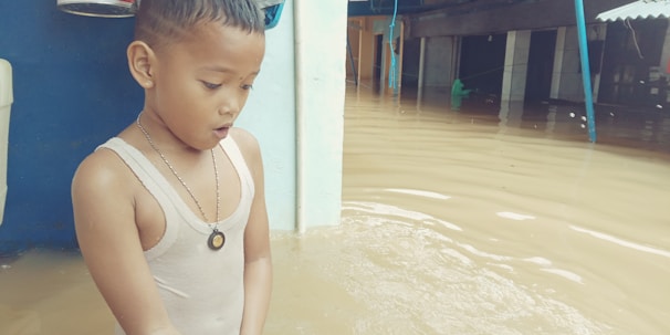 A young child stands in floodwater up to their waist, wearing a sleeveless shirt and necklace. The child looks concerned or surprised. In the background, partially submerged structures and objects are visible.