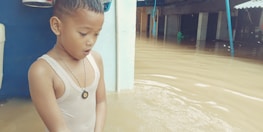 A young child stands in floodwater up to their waist, wearing a sleeveless shirt and necklace. The child looks concerned or surprised. In the background, partially submerged structures and objects are visible.
