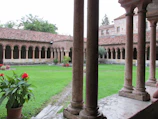 A serene courtyard with arched columns and a neatly maintained grass lawn. The courtyard is surrounded by a cloister with terracotta-colored tiled roofs and lined with evenly spaced columnar arches. Potted plants with red flowers and green foliage are scattered around, adding color to the scene.