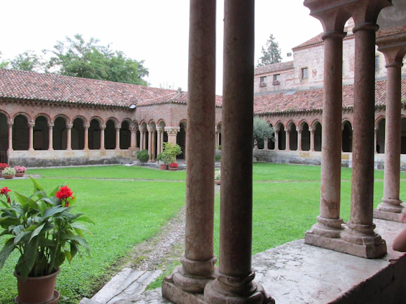 A serene courtyard with arched columns and a neatly maintained grass lawn. The courtyard is surrounded by a cloister with terracotta-colored tiled roofs and lined with evenly spaced columnar arches. Potted plants with red flowers and green foliage are scattered around, adding color to the scene.