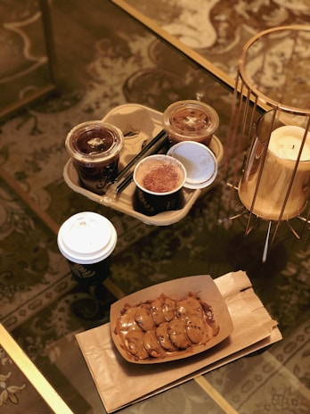 A glass coffee table holds a cardboard tray with two iced coffee cups, one hot coffee cup, and a dessert. Beside the tray is a golden candle holder with a white candle. The table has a decorative patterned carpet underneath.