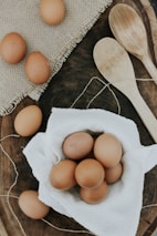 A collection of brown eggs is displayed on a white textured cloth, surrounded by a rustic wooden surface. Two wooden spoons lie nearby, and a portion of a burlap mat adds texture to the scene.