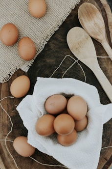 A collection of brown eggs is displayed on a white textured cloth, surrounded by a rustic wooden surface. Two wooden spoons lie nearby, and a portion of a burlap mat adds texture to the scene.