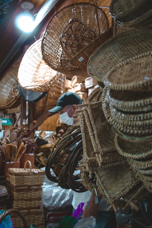 A small shop filled with various woven baskets and wicker crafts hanging from the ceiling and walls. A person wearing a cap and face mask is partially visible, browsing the items. The shop has a warm and cozy atmosphere with soft lighting enhancing the natural tones of the wicker products.