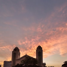 A large church with two tall towers silhouetted against a vibrant sunset sky. The clouds are painted in warm hues of orange and pink, creating a dramatic backdrop. The building's architectural features, including crosses atop the towers, are visible in the silhouette.