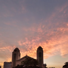A large church with two tall towers silhouetted against a vibrant sunset sky. The clouds are painted in warm hues of orange and pink, creating a dramatic backdrop. The building's architectural features, including crosses atop the towers, are visible in the silhouette.