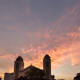A large church with two tall towers silhouetted against a vibrant sunset sky. The clouds are painted in warm hues of orange and pink, creating a dramatic backdrop. The building's architectural features, including crosses atop the towers, are visible in the silhouette.