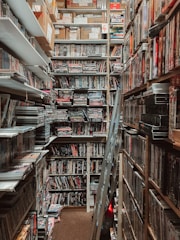 A narrow, cluttered storage room filled with shelves of DVDs and boxes stacked up to the ceiling. A stepladder is located in the middle of the aisle, and the lighting is dim, casting shadows over the items.