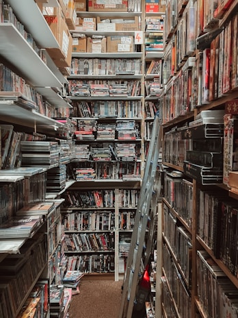 A narrow, cluttered storage room filled with shelves of DVDs and boxes stacked up to the ceiling. A stepladder is located in the middle of the aisle, and the lighting is dim, casting shadows over the items.