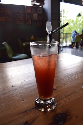 A tall glass of iced tea with a reddish hue sits on a wooden table. The drink is garnished with a straw and a stirrer. The background features a cozy café setting with chairs and wall decorations, and natural light filtering through windows.