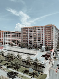 A large residential building with red and white accents rises behind a multi-story parking structure. Palm trees line the edge of the road near the building, with clear skies and clouds above.