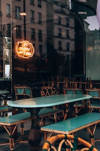 A cozy café interior with rattan chairs and turquoise cushions reflected in a glass window. The reflection includes an urban street view with old buildings. A warm, neon sign reading 'La crème de Paris' adds a touch of elegance.