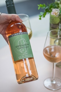 A hand is holding a bottle of rosé wine labeled 'L'Impossible Grenache Cinsault 2019'. Two wine glasses with light pink content are on a table alongside a small vase containing green leaves and a pink flower.