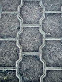 Close-up view of a textured pattern created by interlocking paving stones. The stones have a uniform shape with a consistent texture and subtle color variations.