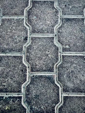 Close-up view of a textured pattern created by interlocking paving stones. The stones have a uniform shape with a consistent texture and subtle color variations.