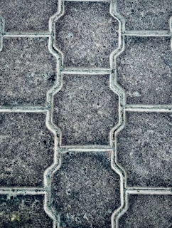 Close-up view of a textured pattern created by interlocking paving stones. The stones have a uniform shape with a consistent texture and subtle color variations.