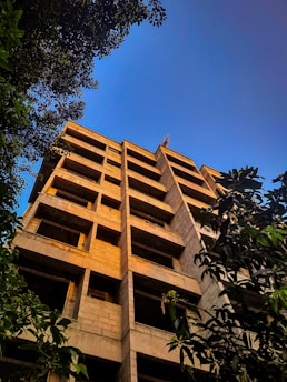 A multi-story building under construction is seen from below, framed by lush green trees. The building has exposed concrete surfaces and no windows or exterior finishes. The bright blue sky in the background adds contrast to the structure.