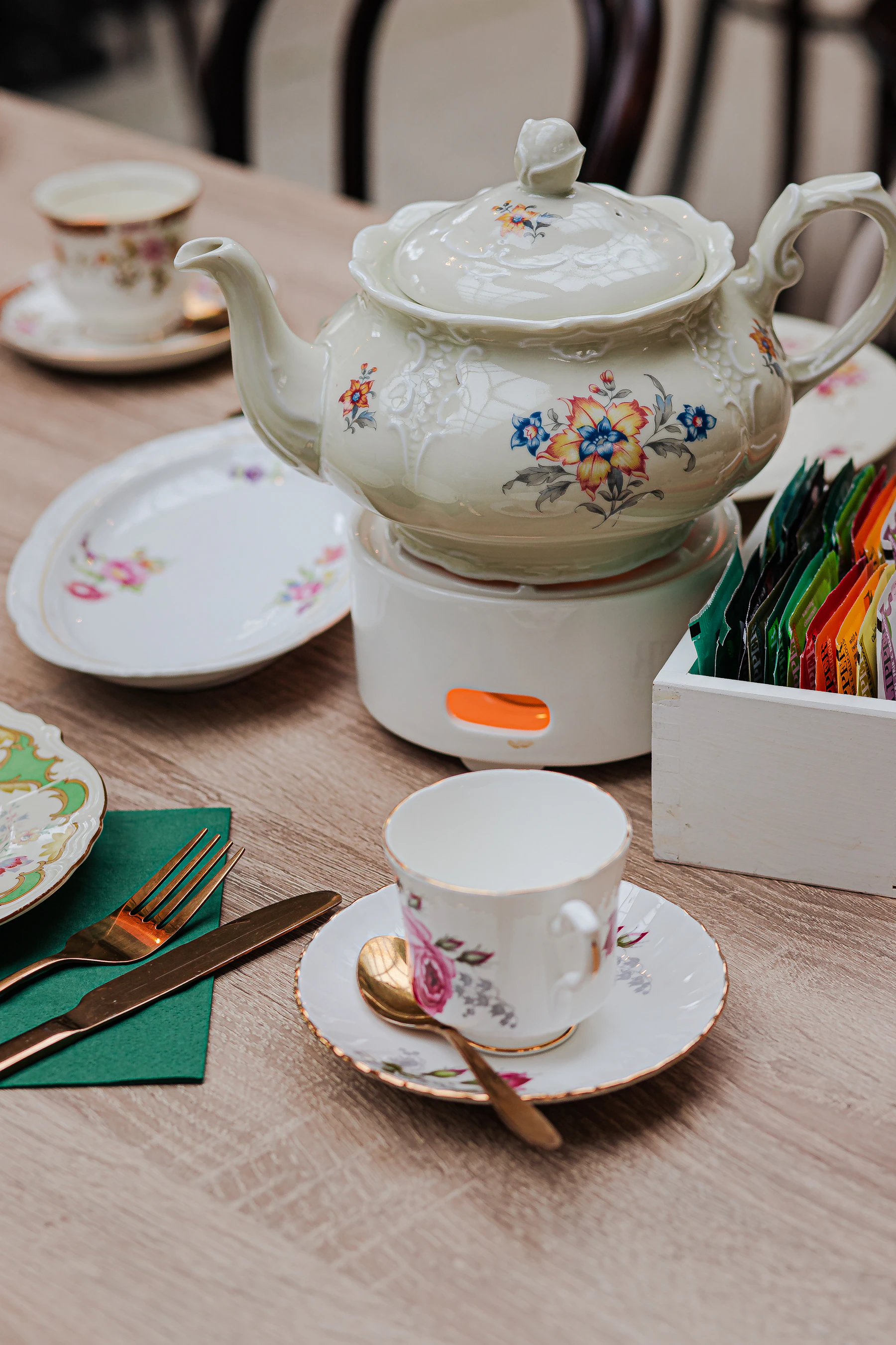 A cream teapot painted with flowers on a warmer, surrounded by floral cups, saucers and plates