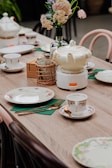 A wooden table is elegantly set with fine china, including a teapot with floral designs, matching cups, saucers, and plates. A vase with light pink flowers adds a touch of nature to the setting. Cutlery is neatly placed on green napkins, and a woven basket holds sugar packets. The overall arrangement suggests a refined and cozy tea setup.