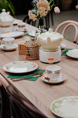 A wooden table is elegantly set with fine china, including a teapot with floral designs, matching cups, saucers, and plates. A vase with light pink flowers adds a touch of nature to the setting. Cutlery is neatly placed on green napkins, and a woven basket holds sugar packets. The overall arrangement suggests a refined and cozy tea setup.
