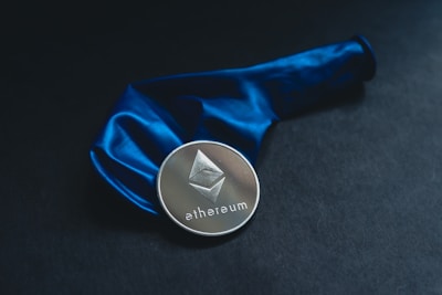 A silver-colored Ethereum coin is placed on a dark surface, next to an inflated blue balloon. The Ethereum logo and text are clearly visible on the coin.