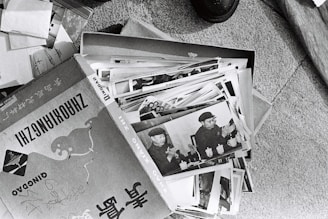 A collection of black and white photographs is spread out, primarily showing an individual in a military uniform. The images appear to be vintage, possibly documenting historical events or figures. The photographs are slightly worn and are placed in a cardboard box, which has text and illustrations on it.