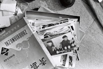 A collection of black and white photographs is spread out, primarily showing an individual in a military uniform. The images appear to be vintage, possibly documenting historical events or figures. The photographs are slightly worn and are placed in a cardboard box, which has text and illustrations on it.