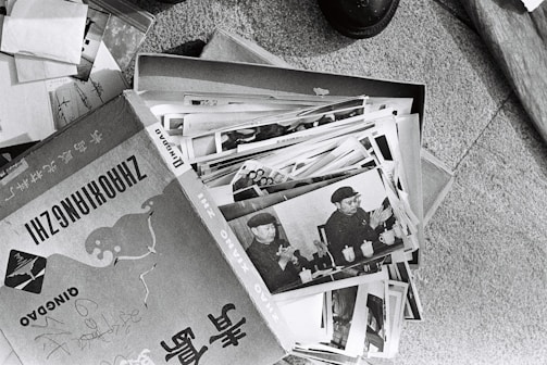 A collection of black and white photographs is spread out, primarily showing an individual in a military uniform. The images appear to be vintage, possibly documenting historical events or figures. The photographs are slightly worn and are placed in a cardboard box, which has text and illustrations on it.