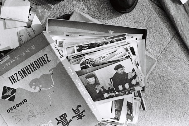 A collection of black and white photographs is spread out, primarily showing an individual in a military uniform. The images appear to be vintage, possibly documenting historical events or figures. The photographs are slightly worn and are placed in a cardboard box, which has text and illustrations on it.