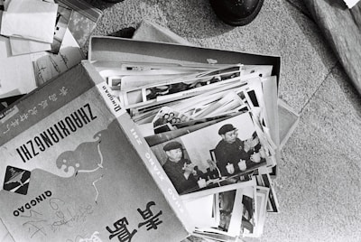 A collection of black and white photographs is spread out, primarily showing an individual in a military uniform. The images appear to be vintage, possibly documenting historical events or figures. The photographs are slightly worn and are placed in a cardboard box, which has text and illustrations on it.
