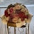 A bouquet of various colored roses and small flowers wrapped in elegant paper. The bouquet features prominently pink and red roses, with accents of white and purple flowers. It is placed on a decorative stand with a soft, blurred background.