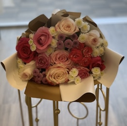 A bouquet of various colored roses and small flowers wrapped in elegant paper. The bouquet features prominently pink and red roses, with accents of white and purple flowers. It is placed on a decorative stand with a soft, blurred background.