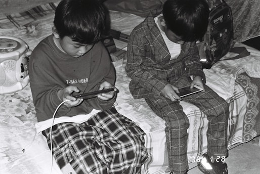 two young boys sitting on a bed looking at their cell phones