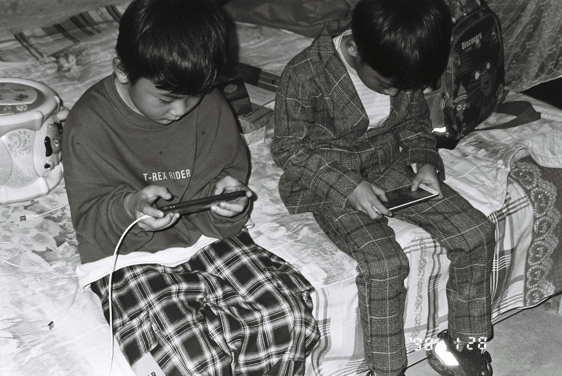 two young boys sitting on a bed looking at their cell phones