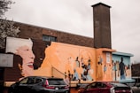A vibrant mural covers the side of a brick building, featuring diverse groups of people interacting and expressing joy. The mural includes large, colorful portraits and scenes promoting community and unity. Several cars are parked below the mural.