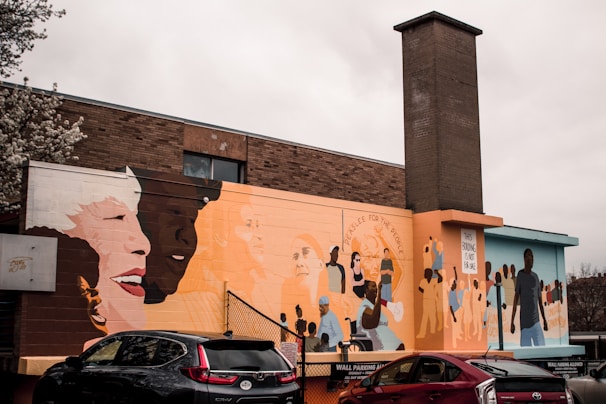 A vibrant mural covers the side of a brick building, featuring diverse groups of people interacting and expressing joy. The mural includes large, colorful portraits and scenes promoting community and unity. Several cars are parked below the mural.