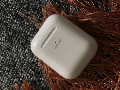 A white charging case labeled 'JOYROOM' is lying on an orange woven fabric placed on a textured, gray carpet.