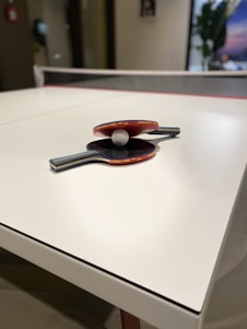A white table tennis table is seen with two rackets and a ball placed on its surface. The rackets have red rubber surfaces and wooden handles. The ball is small and white, positioned between the two rackets. The setting appears to be indoors with a blurred background that includes a plant and a poster.